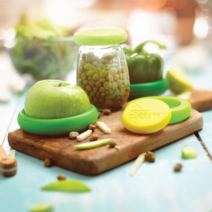 Food huggers cover apple and jar on wooden board, surrounded by green bell pepper, food scraps, and yellow-green lids in a bright kitchen setting.