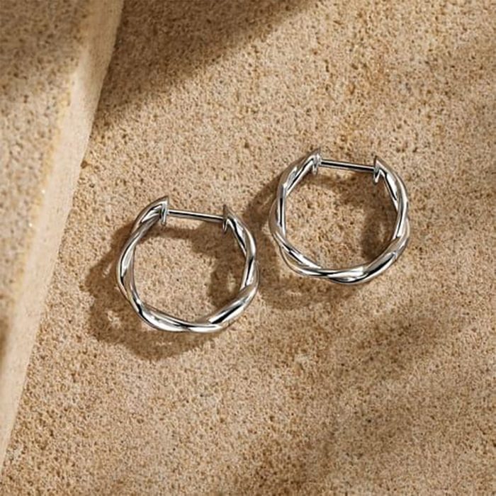 Two silver hoop earrings lie on a textured, sandy surface, casting subtle shadows in natural light.