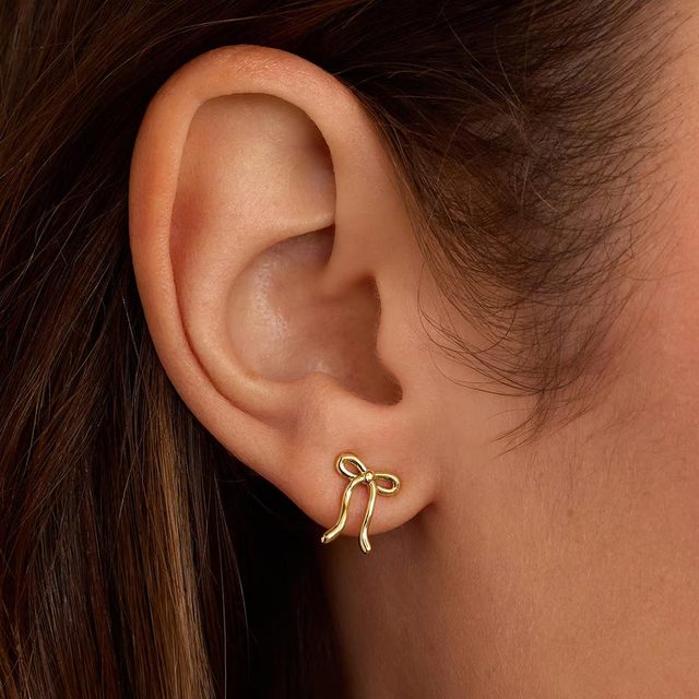 Gold bow-shaped earring adorns an ear, resting against light brown hair in a close-up view.