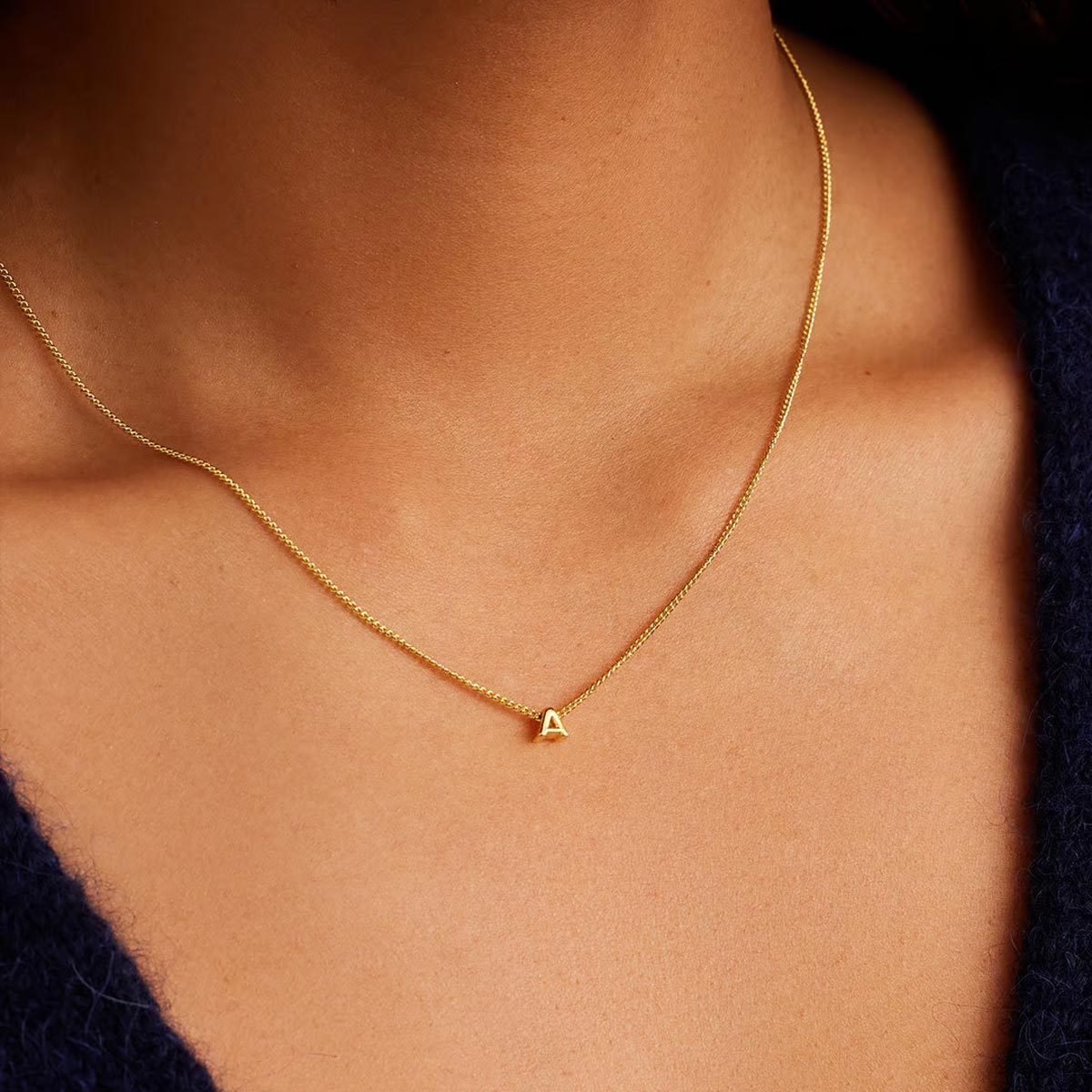 Gold necklace rests on skin, featuring a small letter "A" charm, surrounded by dark blue fabric.