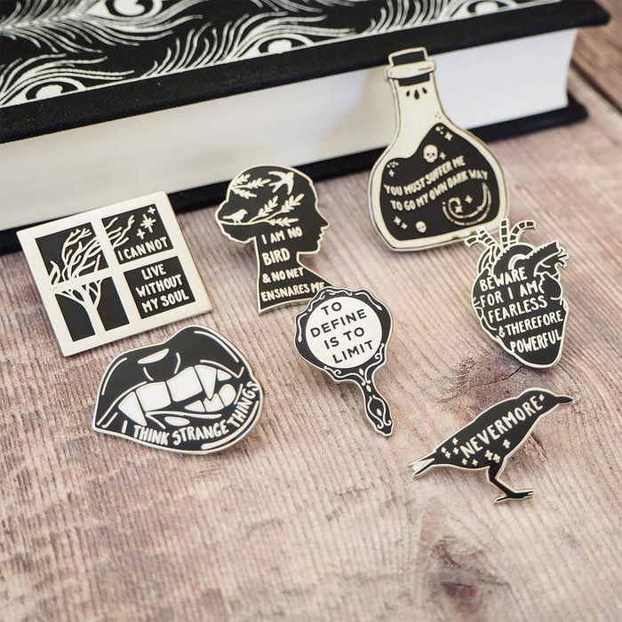 Enamel pins display literary quotes on a wooden surface beside a patterned book.
