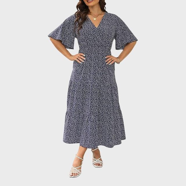 A woman models a navy floral dress with short sleeves and a V-neck, standing against a plain background, wearing white strappy sandals.