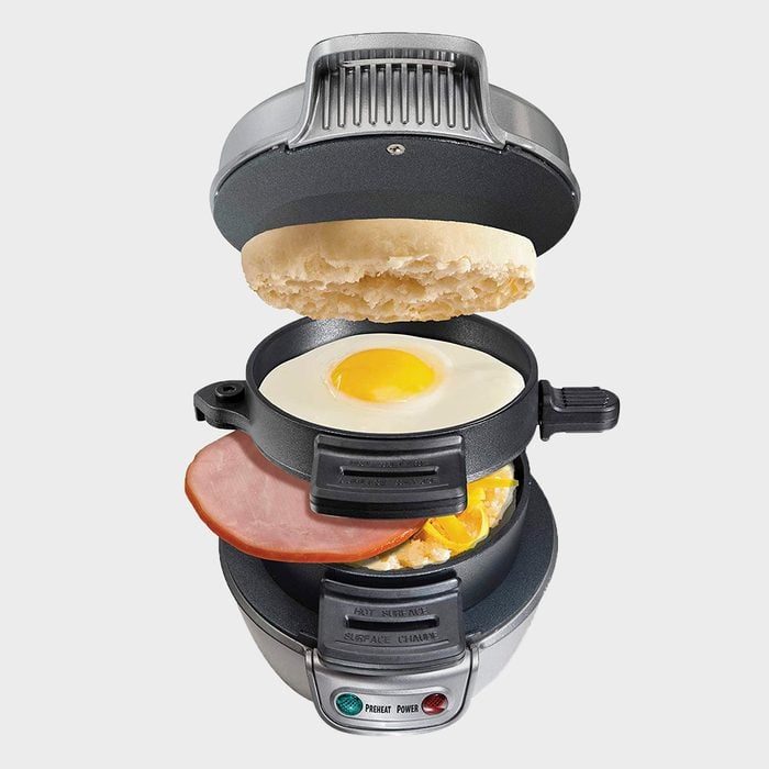 Sandwich maker assembling egg, ham, cheese, and muffin layers; positioned open with ingredients floating in layers, set against a neutral background.