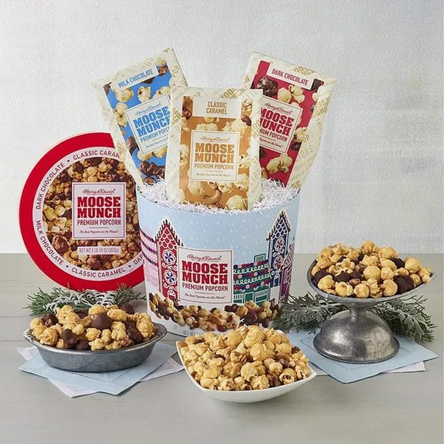 Popcorn bags displayed in a decorative tin, surrounded by bowls of popcorn on a table with festive greenery. Packaging reads 