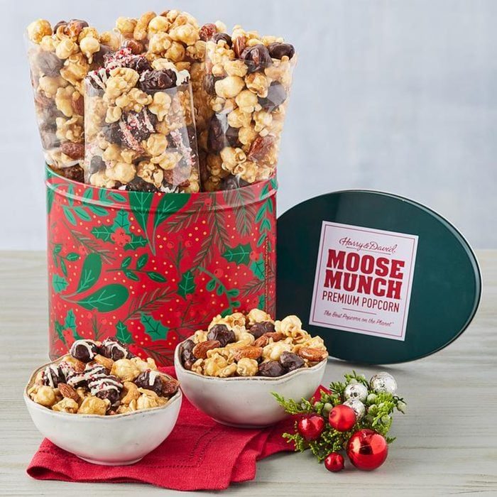 Popcorn packaged and displayed in a festive red tin is paired with two bowls, set on a red cloth with holiday decorations around.