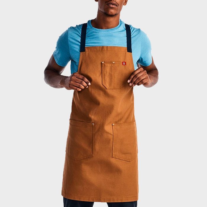 Apron hangs over blue-shirted person's chest, hands holding edges, against plain background. Three pockets visible, a small red label on top pocket.