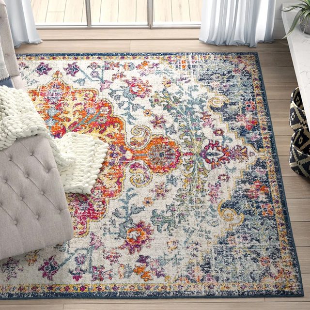 A colorful, patterned rug lies on a wooden floor beside a tufted sofa and a knitted white blanket near large windows and curtains.