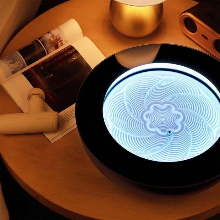 Lit kinetic sand sculpture creates intricate patterns on tabletop near books, massage gun, and a speaker, under warm ambient lighting.