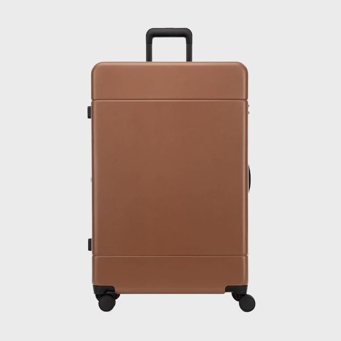 Brown suitcase stands upright with four wheels; black telescopic handle extends at the top. Neutral background highlights the minimalist design.