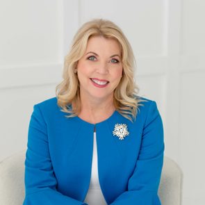 A woman is smiling, sitting on a chair, wearing a blue jacket with a decorative brooch, against a plain white background.
