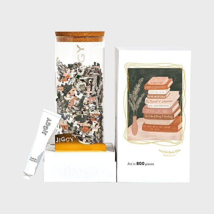 Jigsaw puzzle pieces filling a clear, rectangular container alongside a box showing stacked books illustration; set includes a tube labeled 