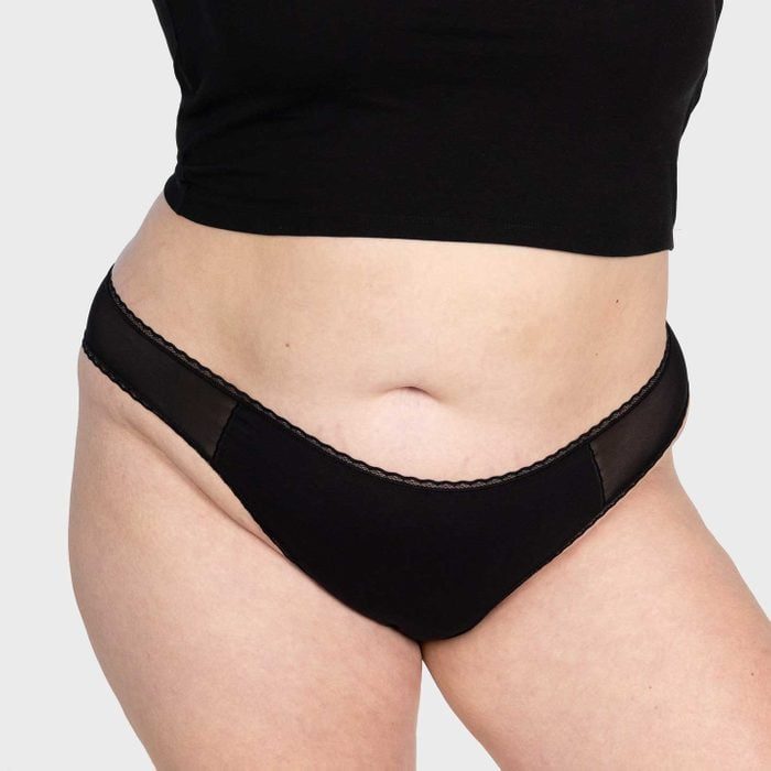 A person wears black underwear, standing with a cropped black top, against a plain light background.