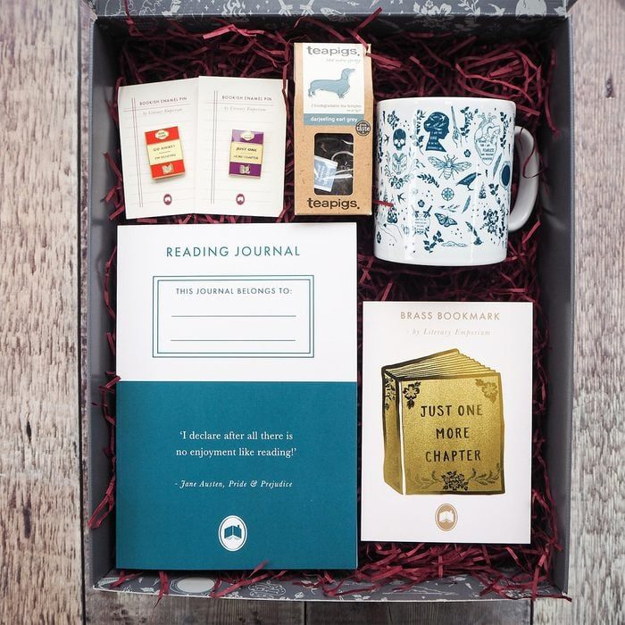 Box contains a mug, reading journal, teabags, pins, and brass bookmark, all arranged on red shredded paper.