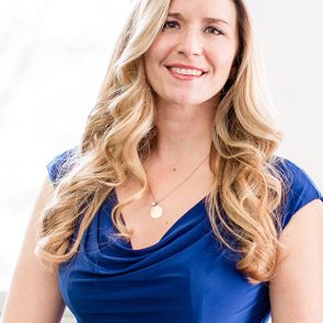 A woman smiles confidently, wearing a blue dress and necklace, standing in a softly lit indoor setting.