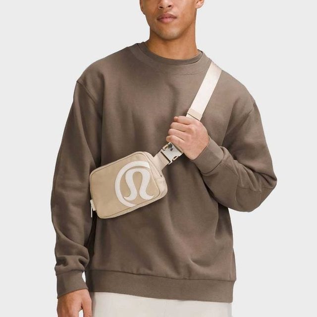 A person wears a beige crossbody bag with a logo, standing in a neutral setting, wearing a brown sweatshirt.
