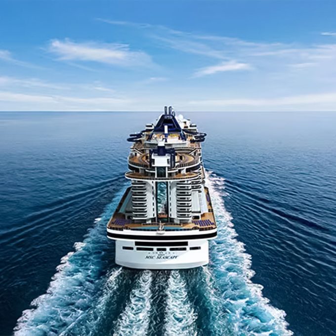 Cruise ship sails forward, leaving a wide, foamy wake, surrounded by open, calm ocean under a clear blue sky.