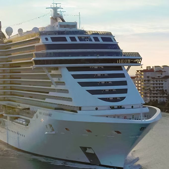 Cruise ship sails through a calm harbor, passing by waterfront buildings under a bright, clear sky.