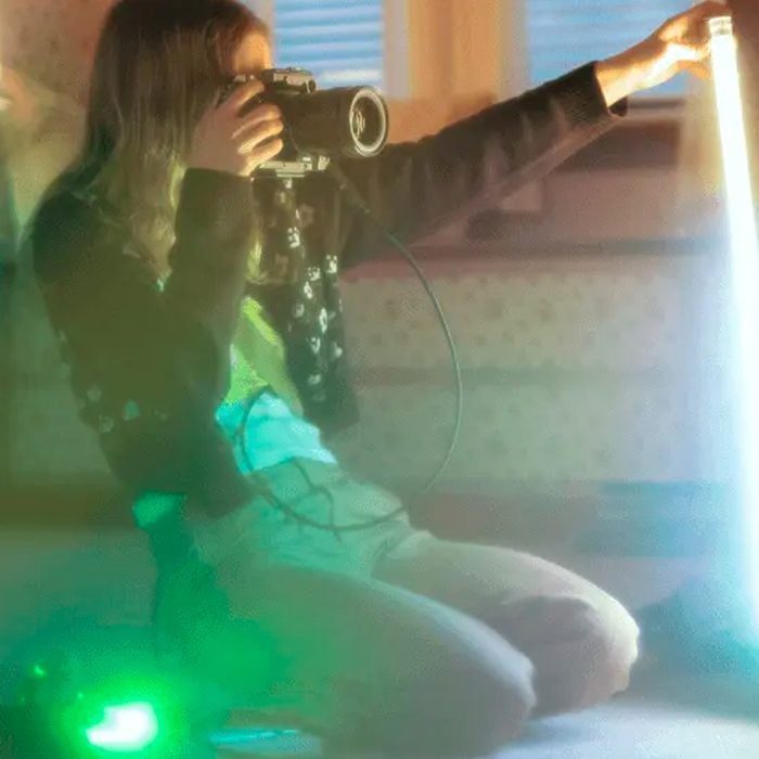 Person kneeling holds a camera, photographing a glowing light tube in a softly lit room.