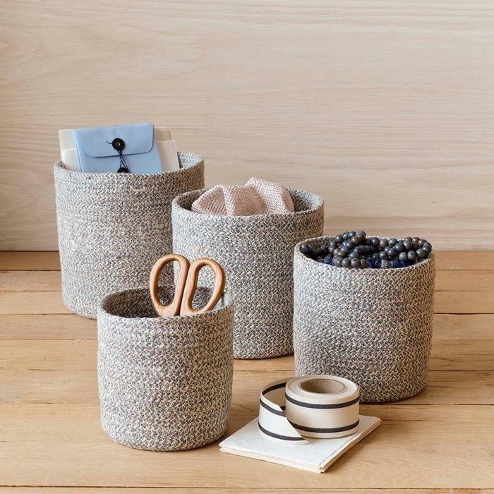 Gray woven baskets hold various items like letters, cloth, beads, and scissors on a wooden surface, accompanied by rolls of ribbon and paper.