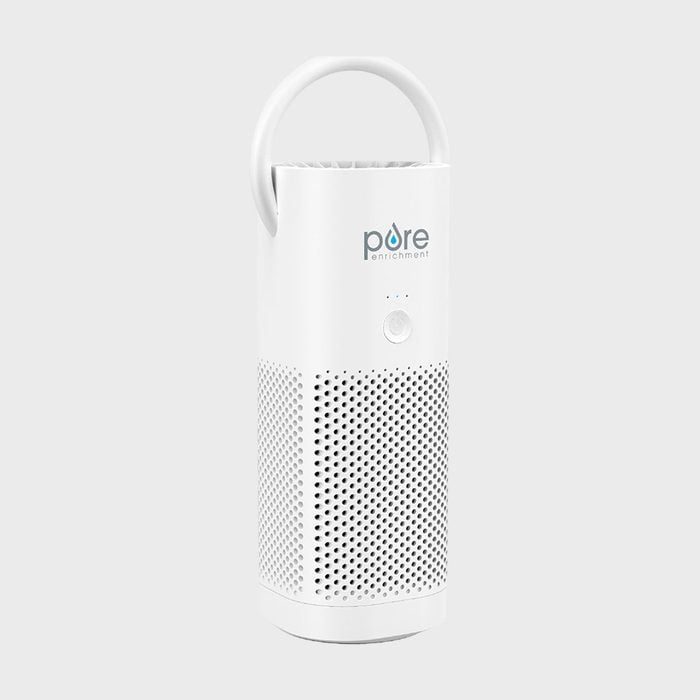 A white cylindrical air purifier stands upright, featuring a handle and perforated surface, set against a plain gray background. Text: 