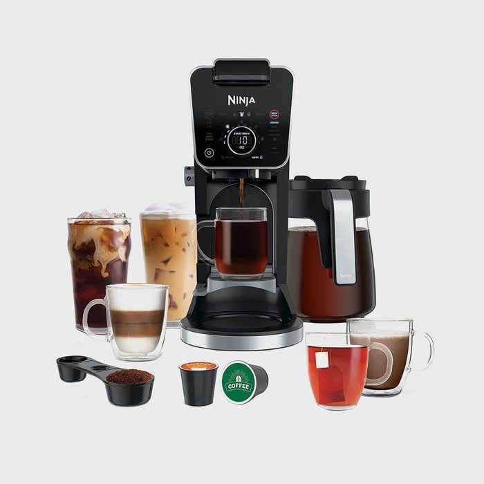 Coffee maker brews various drinks; surrounded by cups of iced coffee, cappuccino, hot tea, and pods; black carafe nearby.