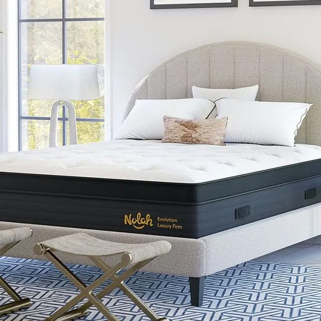 Mattress resting on a bed frame with pillows, in a bright room with a window and geometric-patterned rug. Text: 