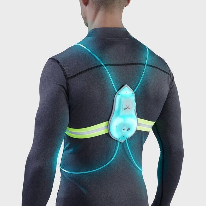 Man wearing illuminated harness, standing against plain background; the harness emits a blue glow and features a bright yellow horizontal strap.