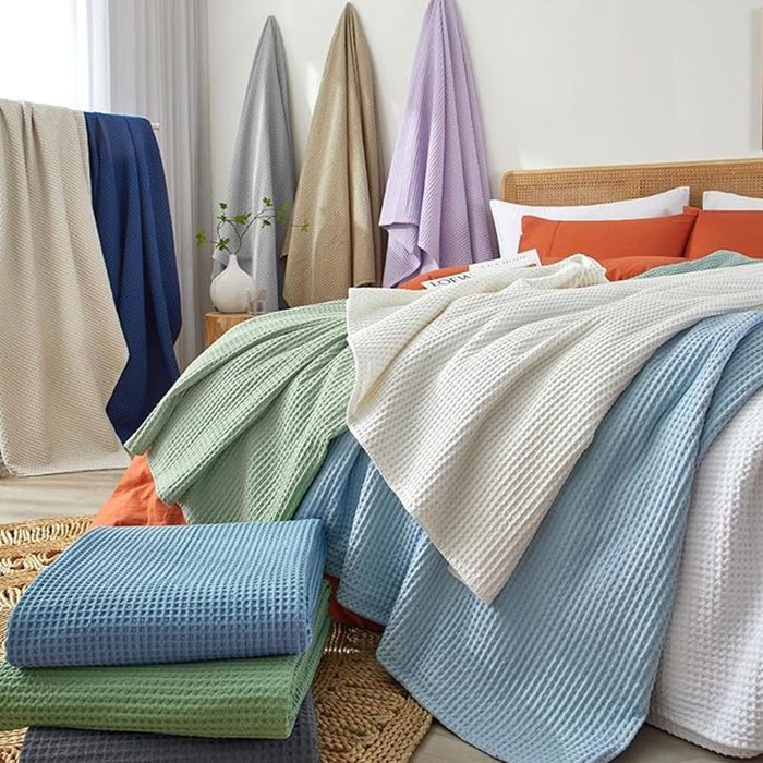Blankets drape across a bed; multi-colored fabrics hang on walls. Soft lighting highlights the neatly arranged, vibrant textiles in a cozy bedroom setting.