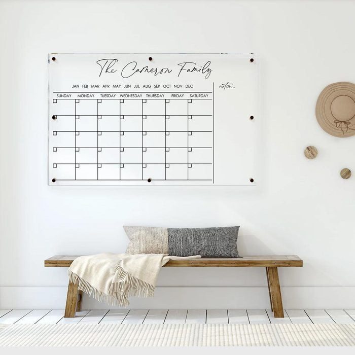 Calendar hangs on wall displaying grid layout. Below, a wooden bench with cushions and a blanket sits on a tiled floor.