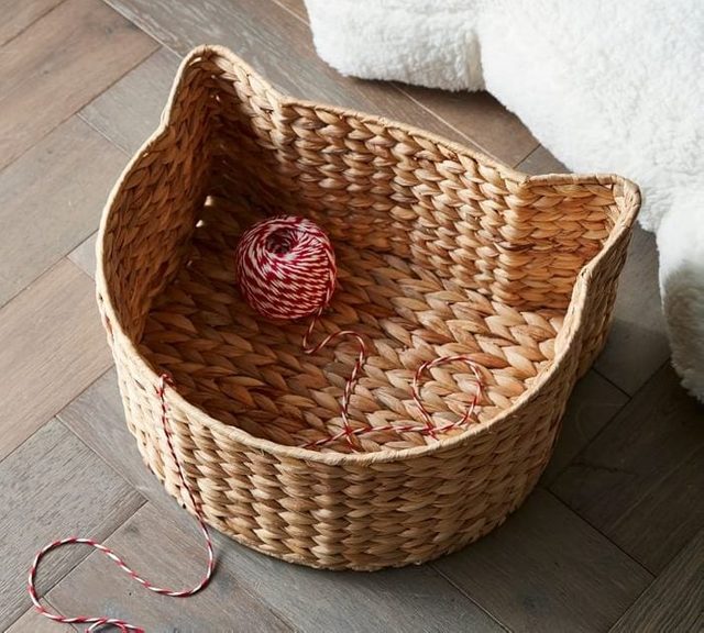 Woven basket shaped like a cat's head holds red and white twine ball, on a wooden floor near a fluffy object.