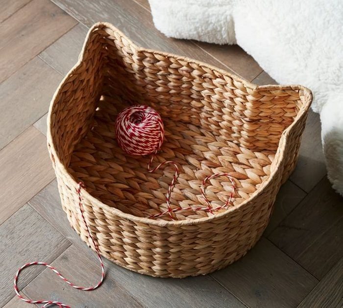 Woven basket shaped like a cat's head holds red and white twine ball, on a wooden floor near a fluffy object.