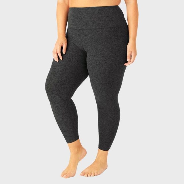 Barefoot person in dark leggings stands against a plain gray background, showing the leggings' fit and high waistband.
