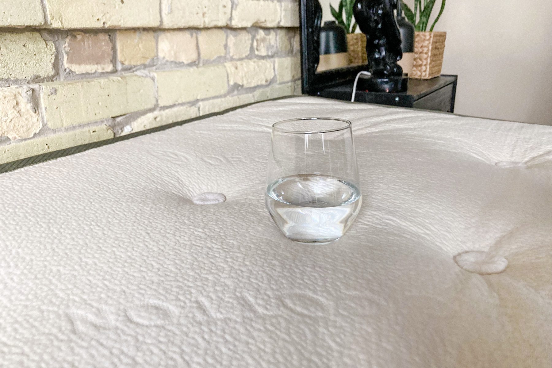 Glass of water resting on a textured mattress surface in a room with a brick wall and potted plants nearby.