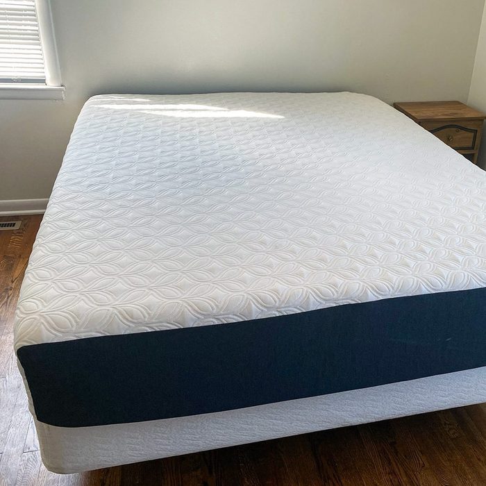 A neatly made bed with a white patterned mattress is in a room with wooden floors and a small wooden nightstand beside it.