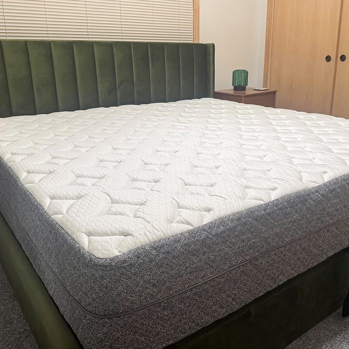 Bed with a thick, white-patterned mattress sits in a bedroom with a dark green headboard and wood furniture, under closed blinds.