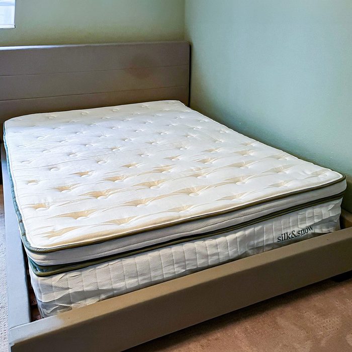 Mattress resting on a brown frame, labeled â€œsilk & snow,â€ in a room with green walls and a small window.