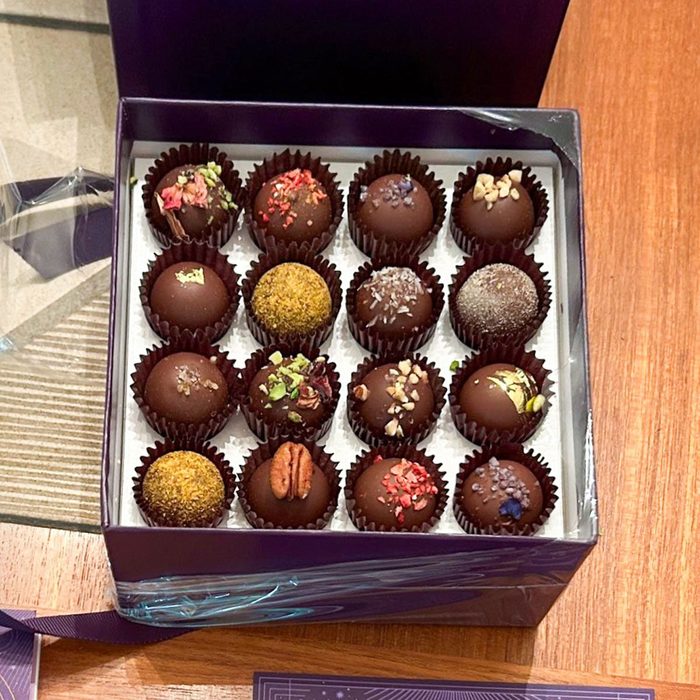 Box of assorted chocolate truffles rests open, each delicately decorated with nuts and sprinkles, displayed on a wooden table surface.