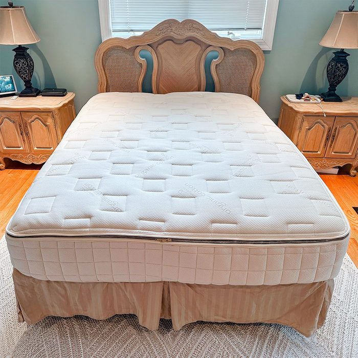 Mattress lies on a bed with wooden headboard, flanked by nightstands, lamps, and window above, in a warmly lit bedroom.