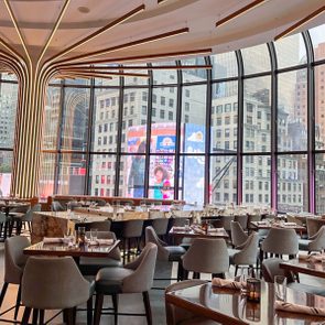 Tables are arranged elegantly in a modern restaurant with large windows overlooking city skyscrapers, featuring visible signs outside reading 