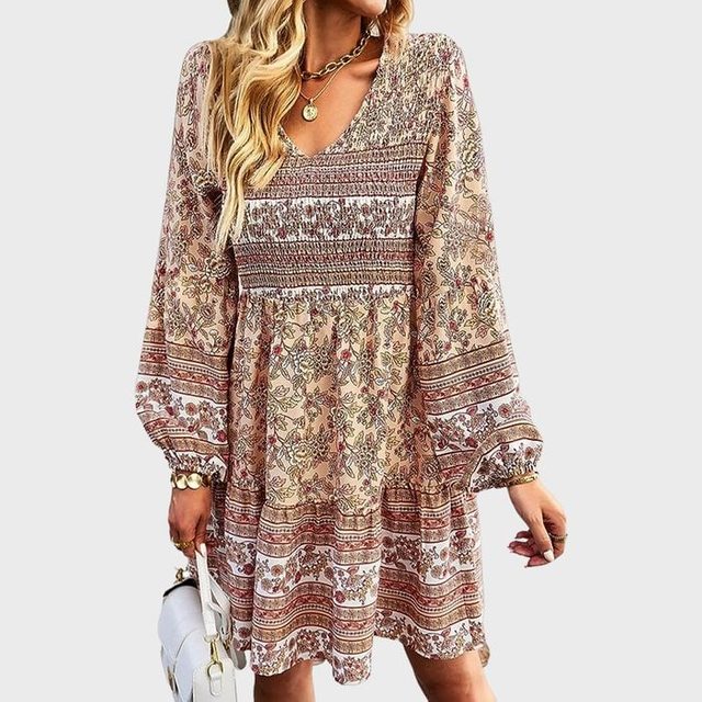 A woman wears a bohemian-style, floral-patterned dress, holding a white handbag in a neutral background.