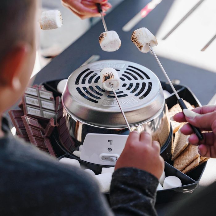 People toast marshmallows over a tabletop grill surrounded by chocolate bars and graham crackers, preparing for making s'mores.
