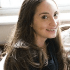 A person with long hair smiling, sitting indoors near a window with soft light filtering through the glass.