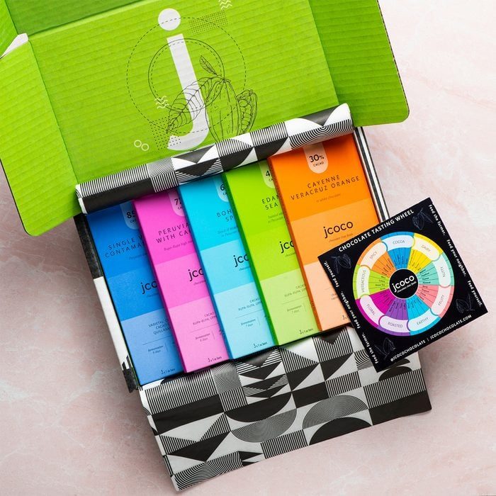 Colorful chocolate bars arranged in an open green box with geometric patterns; a circular tasting wheel sits beside it on a pale pink surface.