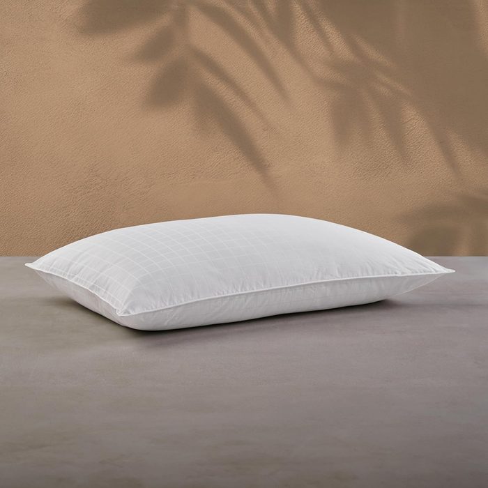 A white pillow rests on a gray surface, with soft, shadowy plant patterns on a beige wall behind it.