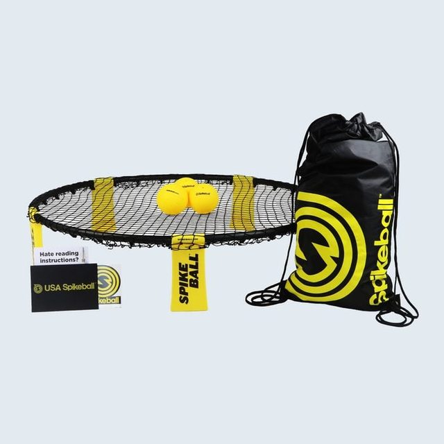 Round net holds two yellow balls, set beside a branded black drawstring bag and instruction booklet, in a Spikeball game setup.