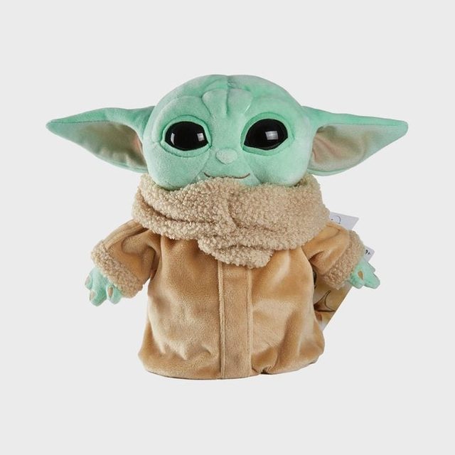 A green plush toy stands wearing a brown robe and scarf, displaying large eyes and prominent ears against a plain background.
