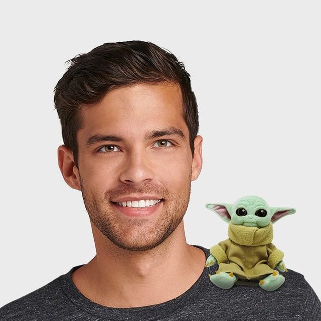 A man smiles with a small, green, alien-like plush toy on his shoulder; both are in front of a plain gray background.