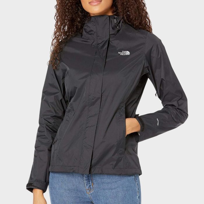 Woman wearing a black jacket stands casually, hand in pocket, against a plain background. The jacket's logo reads 