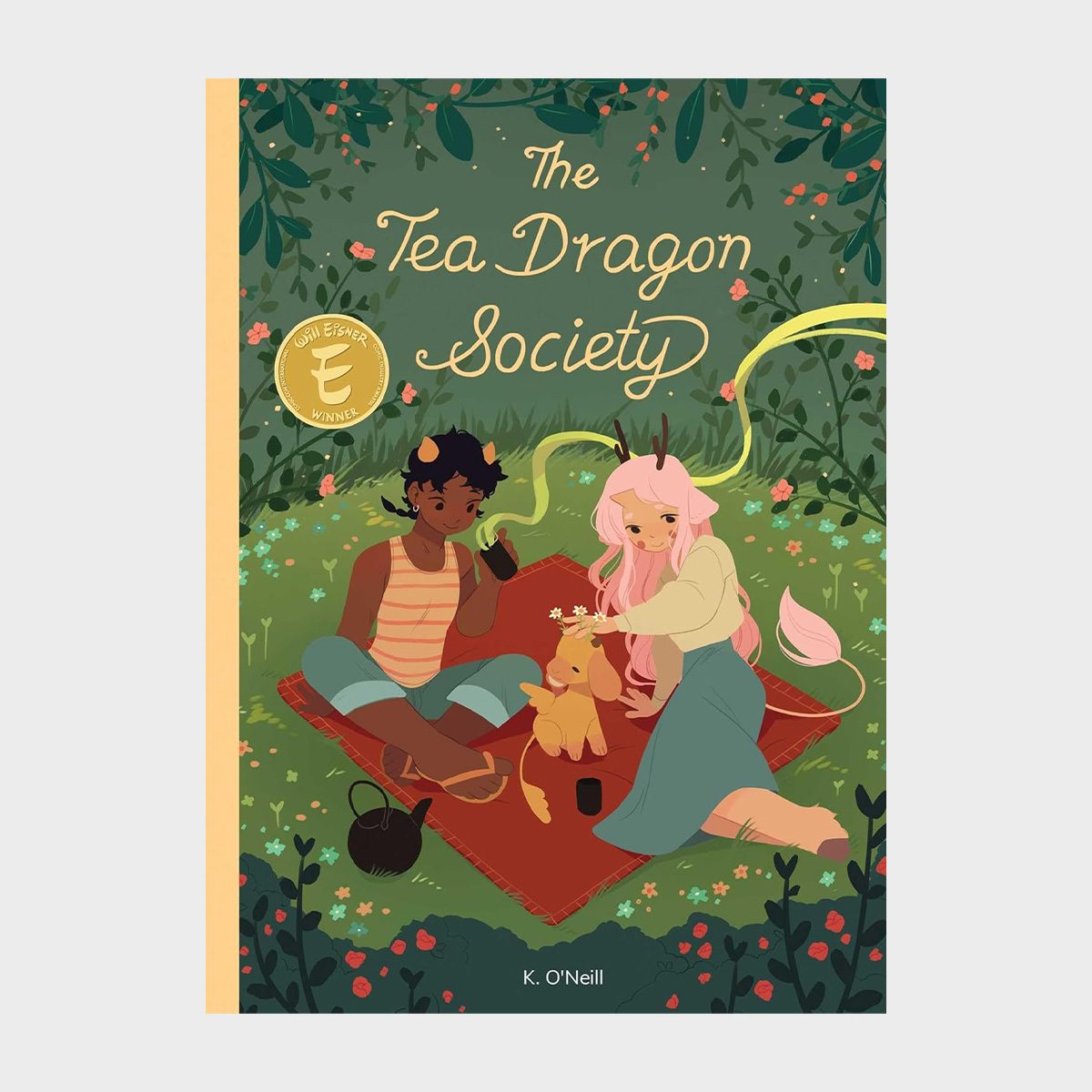 Two characters sit on a picnic blanket with a small dragon-like creature, surrounded by greenery. Text: "The Tea Dragon Society" by K. O'Neill.