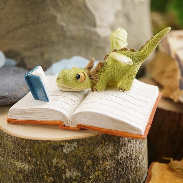 A small green dragon reads a tiny book while lying on an open notebook placed on a wooden surface outdoors.